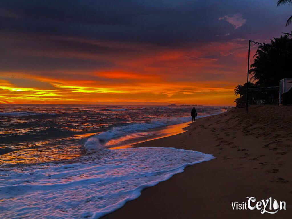 A tranquil evening scene at Hikkaduwa Beach, with the sun setting over the horizon and casting warm hues across the shore.